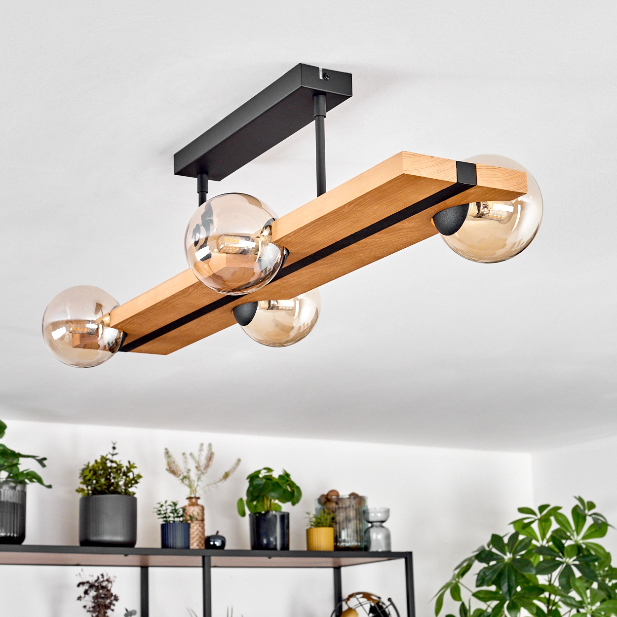 Eine moderne Deckenlampe mit vier Glaskugeln hängt über einem Regal mit Pflanzen und Dekorationen. Die Lampe hat einen Holzbalken.