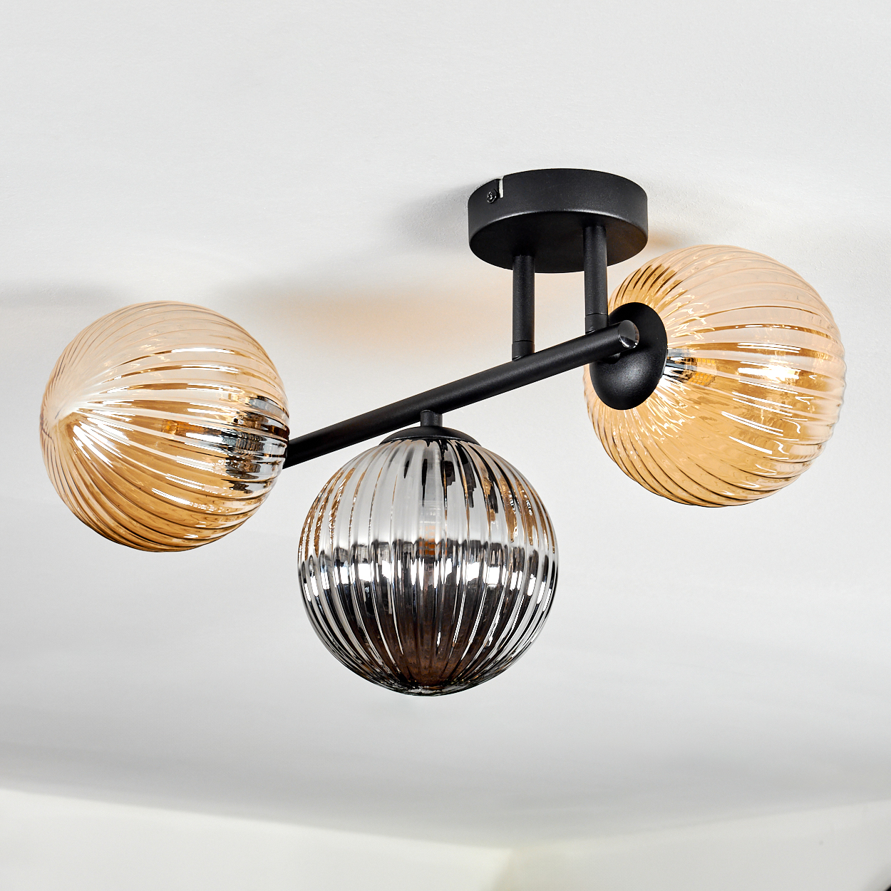 Moderne Deckenleuchte mit drei kugelförmigen Lampen. Zwei sind bernsteinfarben, eine versilbert. Schwarzer Rahmen an weißer Decke.