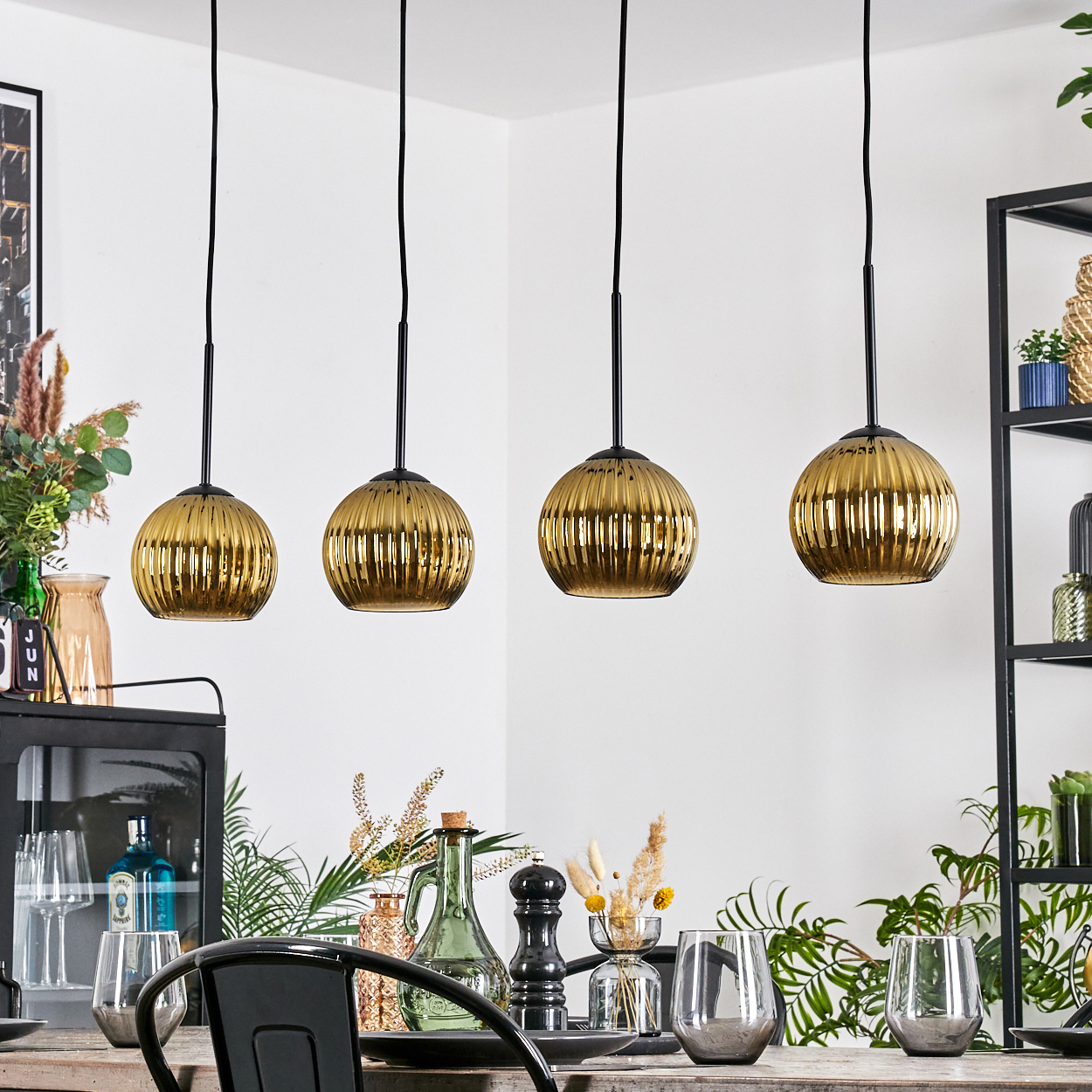 Esszimmer mit vier goldenen Pendelleuchten. Tisch mit Gläsern und Dekor. Pflanzen im Hintergrund, schwarzes Metallregal.