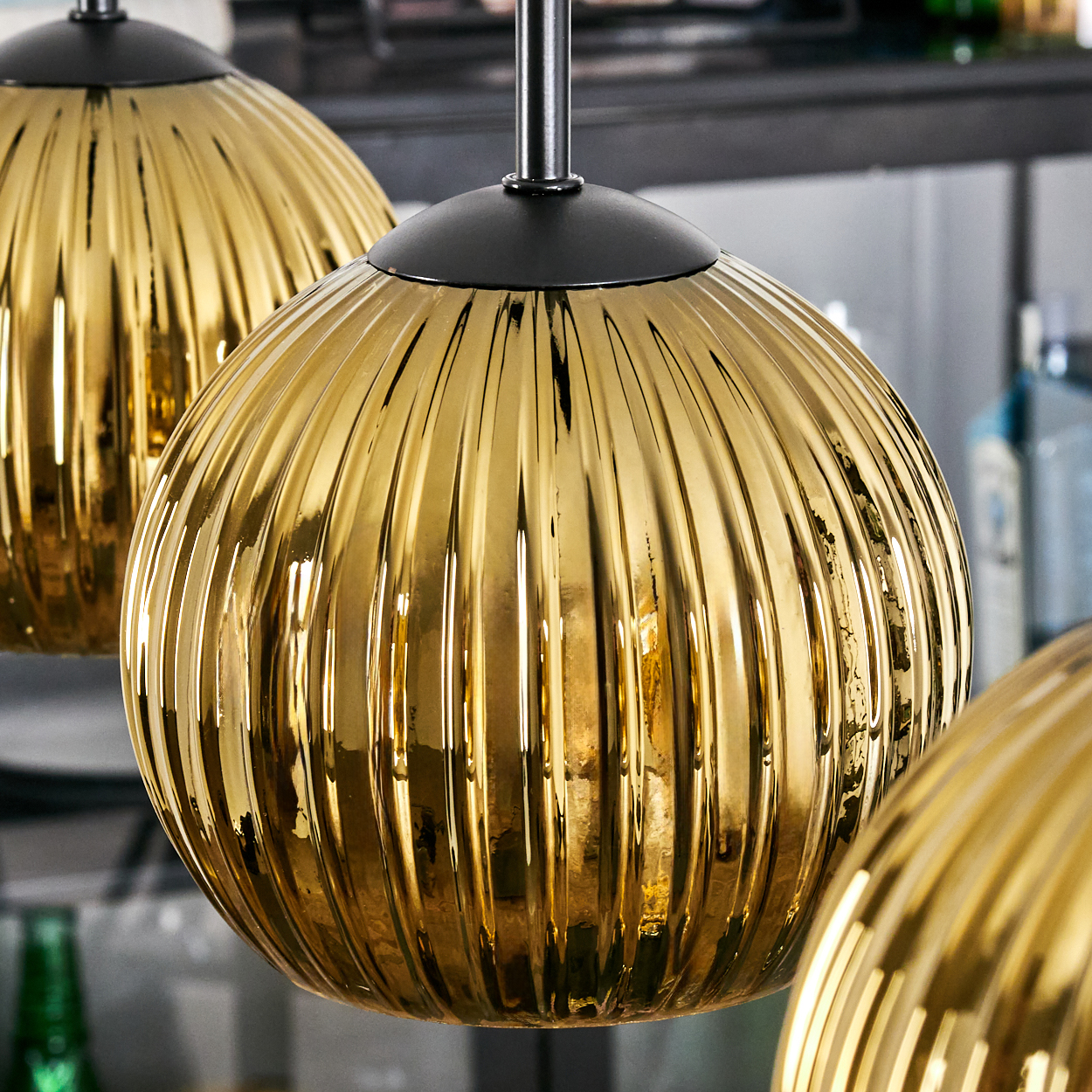 Nahaufnahme von drei goldenen, gerippten Pendelleuchten mit schwarzen Kappen. Im Hintergrund sind ein Regal und Flaschen zu sehen.