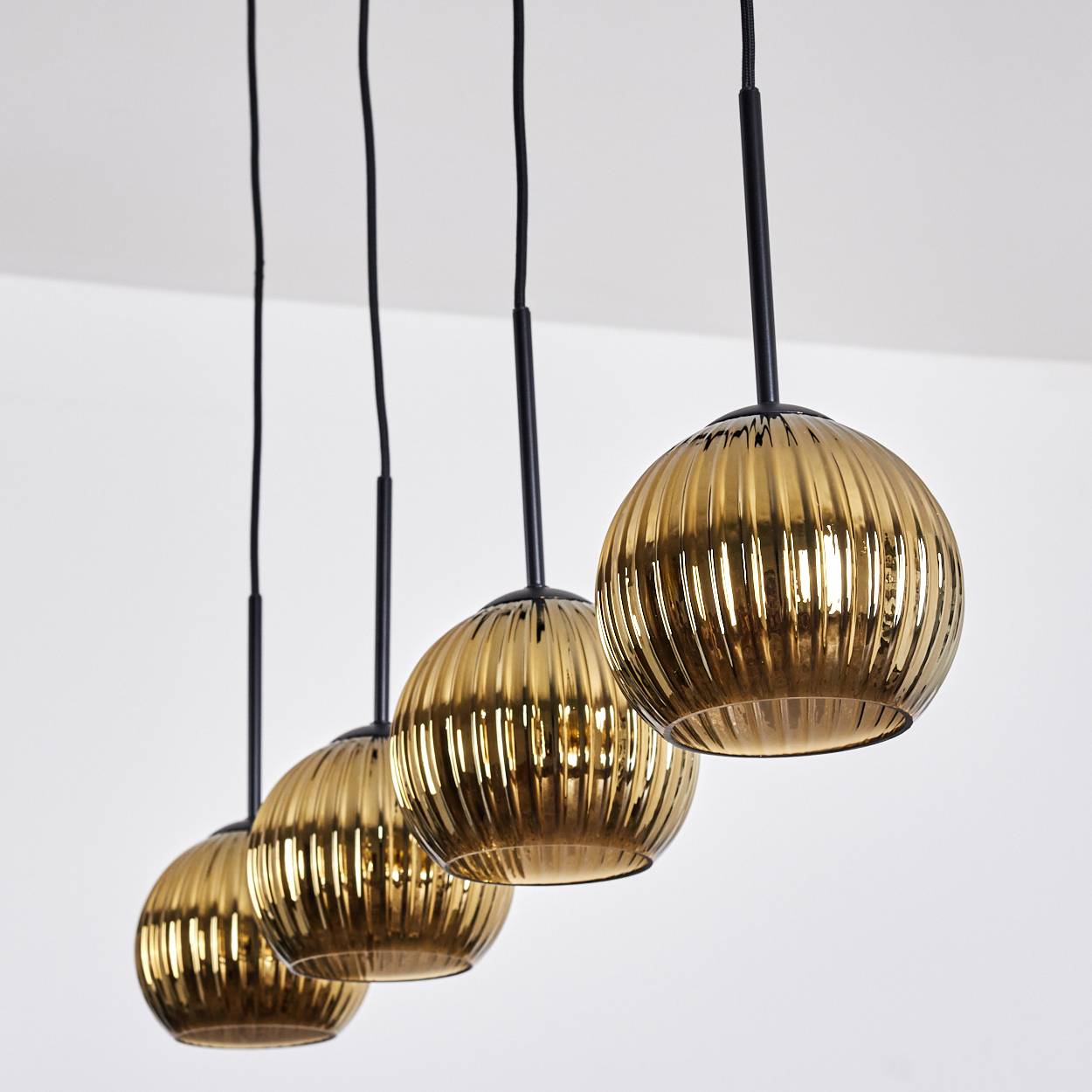 Vier goldene, geriffelte Glas-Pendelleuchten hängen an schwarzen Schnüren und Stangen von einer weißen Decke. Die Leuchten sind rund.