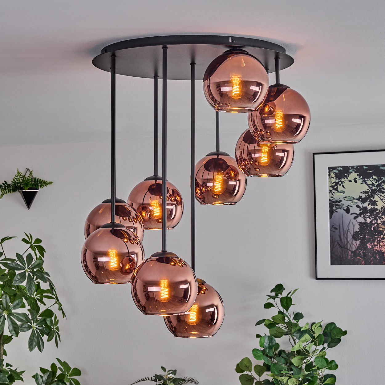 Eine moderne Deckenleuchte mit acht kupferfarbenen Kugellampen. Schwarze Stäbe halten die Lichter, Pflanzen und ein gerahmtes Bild sind im Hintergrund.