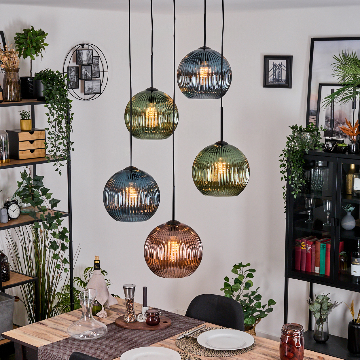 Esszimmer mit Holztisch, Hängelampen und Regalen mit Pflanzen und Dekoration. Teller und eine Karaffe sind auf dem Tisch gedeckt.