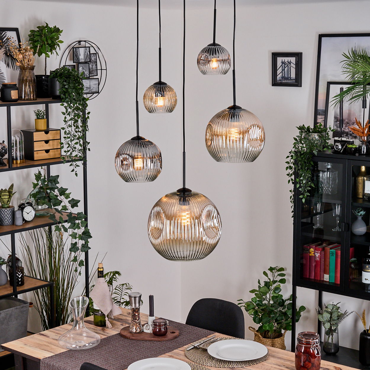 Esszimmer mit Pendelleuchten, Tisch mit Dekoration und Essen, Regale mit Pflanzen und gerahmten Bildern.