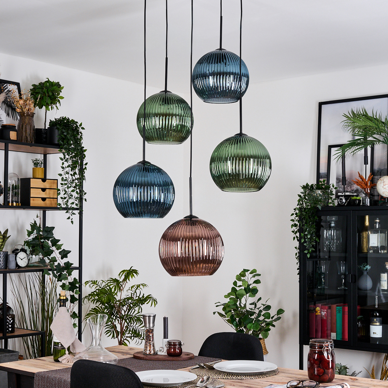 Esszimmer mit Hängelampen. Verschiedene Pflanzen und eine Tischdekoration sind sichtbar. Ein schwarzes Regal und ein Schrank sind im Hintergrund.