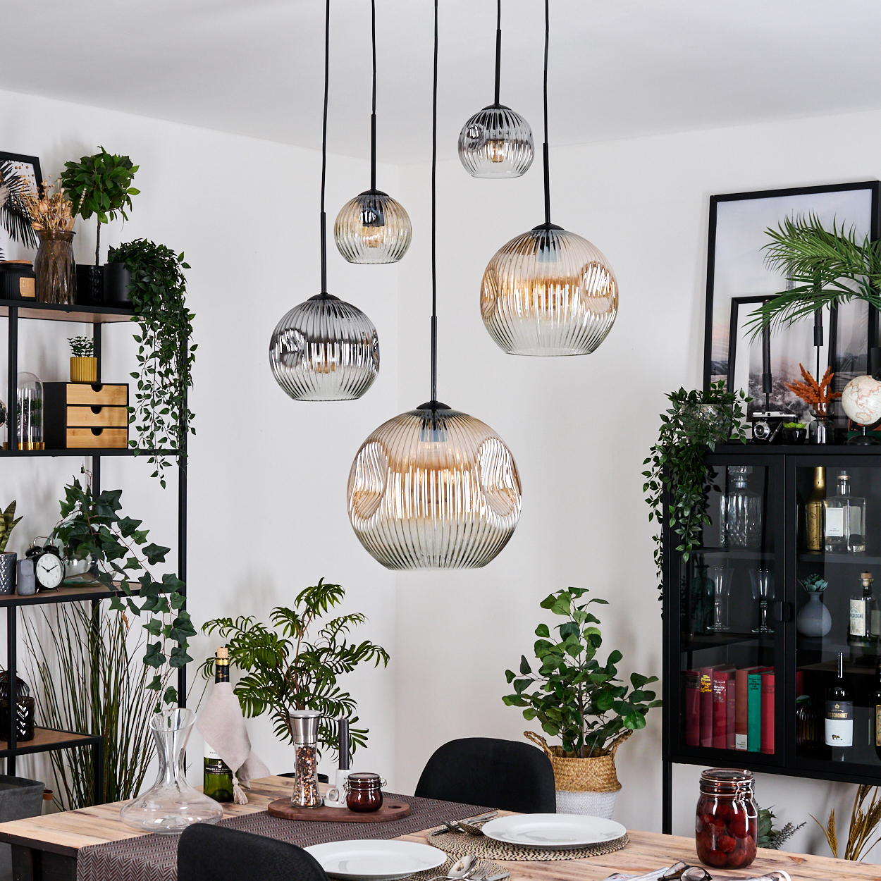 Esszimmer mit hängenden Kugelleuchten. Tisch gedeckt mit Tellern, Pflanzen und schwarzen Regalen mit Dekor.