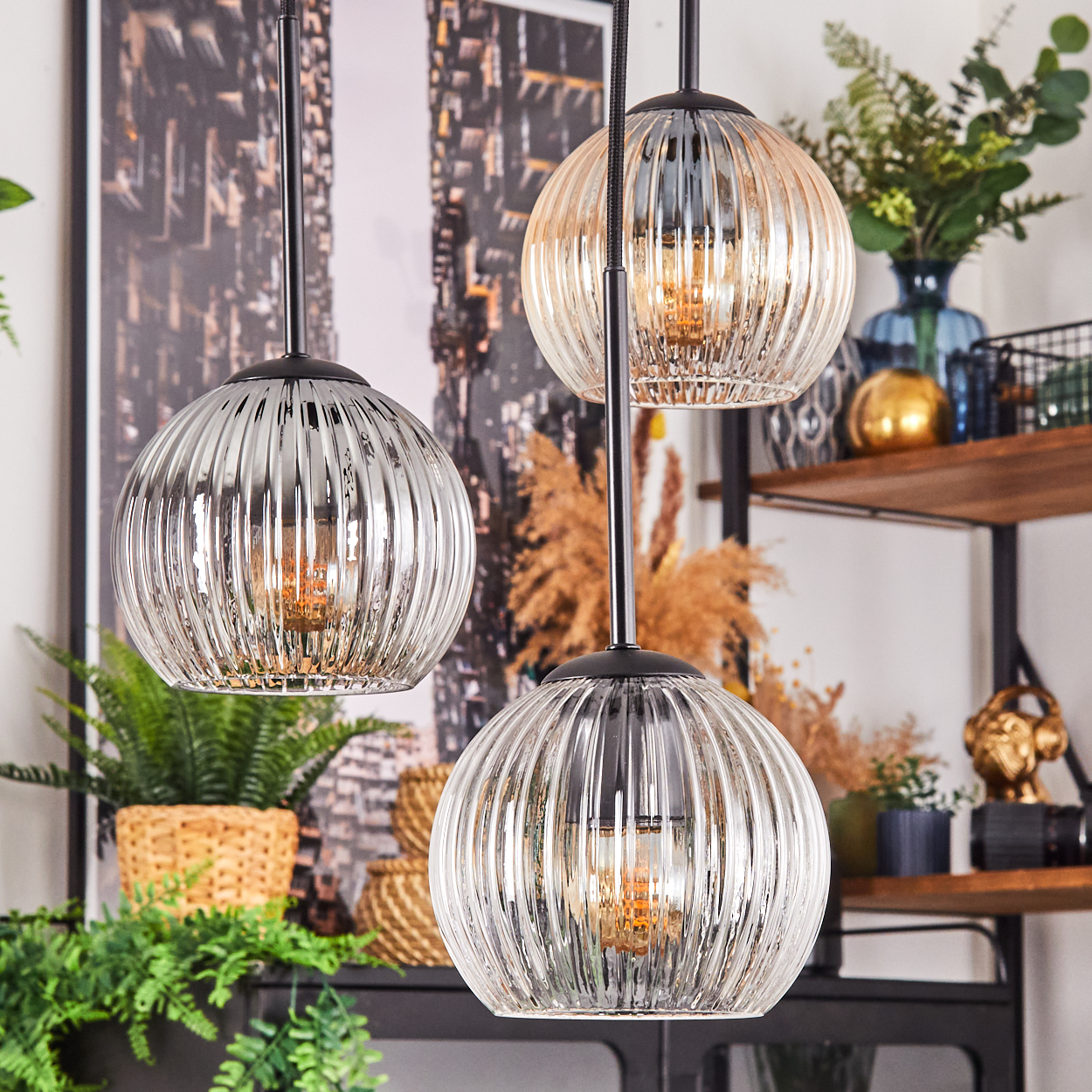 Drei Glas-Pendelleuchten mit geripptem Design hängen in einem Raum. Pflanzen und Dekorationsgegenstände sind im Hintergrund.