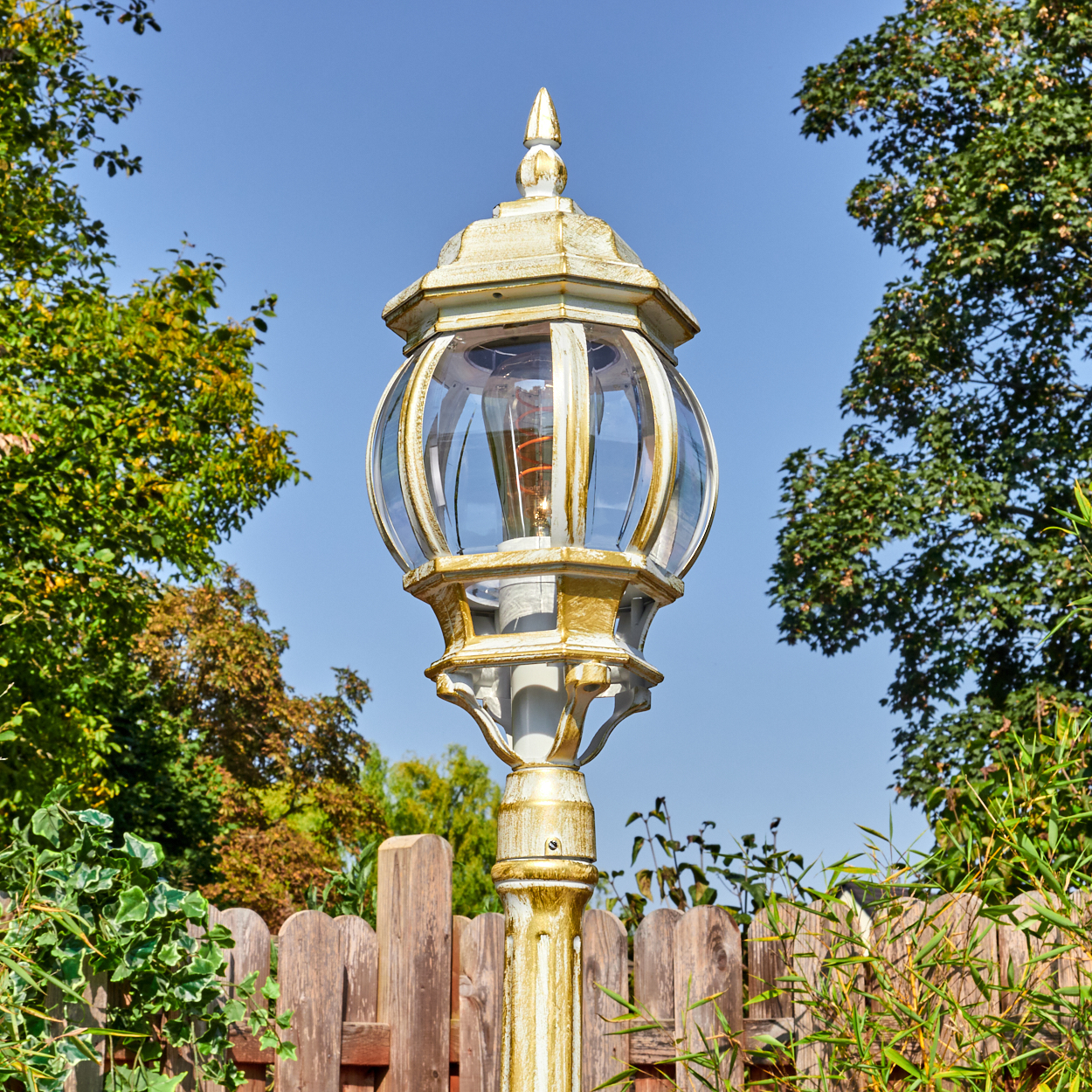 Eine dekorative Außenlampe mit Glaskugel und goldenen Akzenten, vor blauem Himmel, Bäumen und Holzzaun.