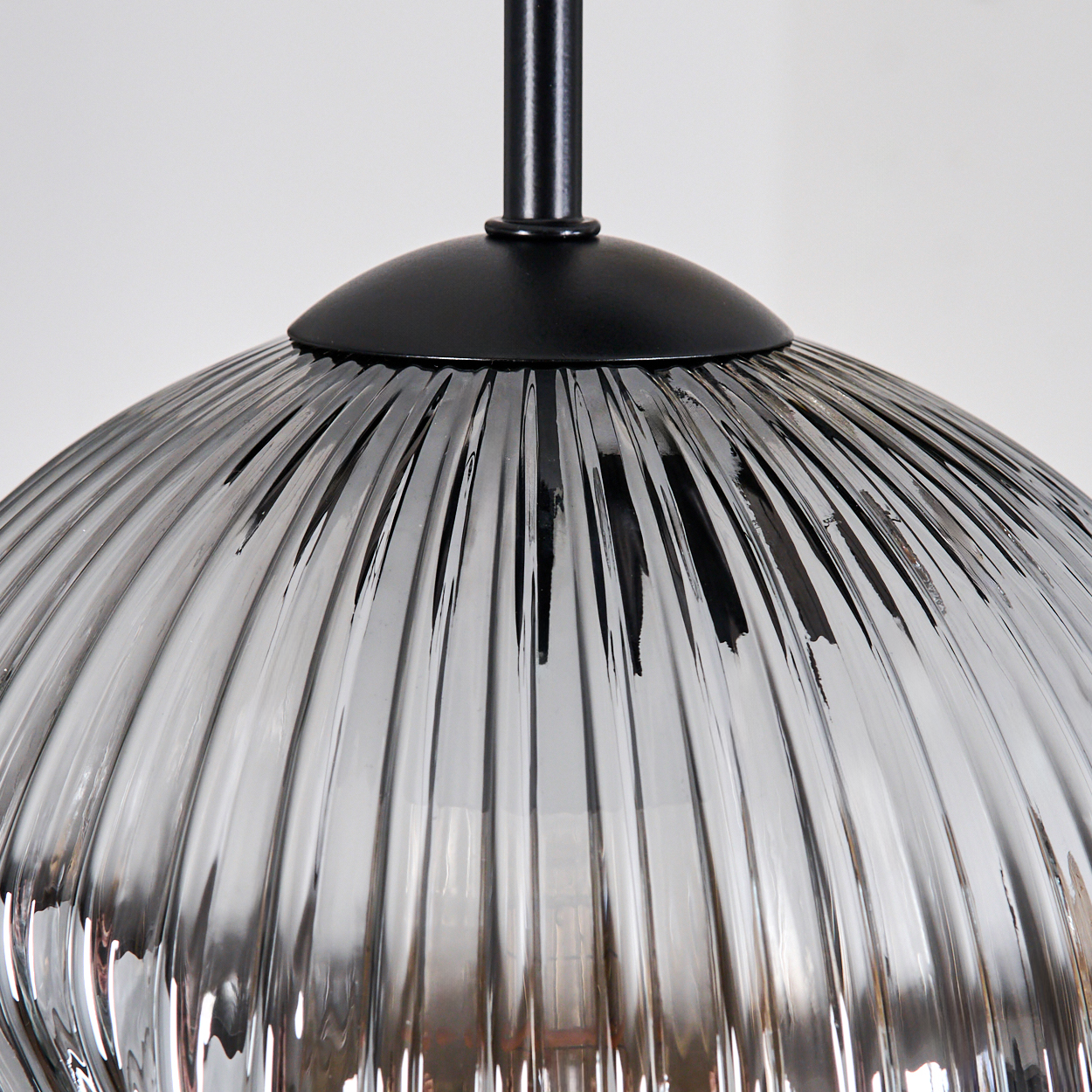 Nahaufnahme eines Lampenschirms aus Glas mit vertikalen Rippen. Er ist grau und hängt an einer schwarzen Metallhalterung vor weißem Hintergrund.