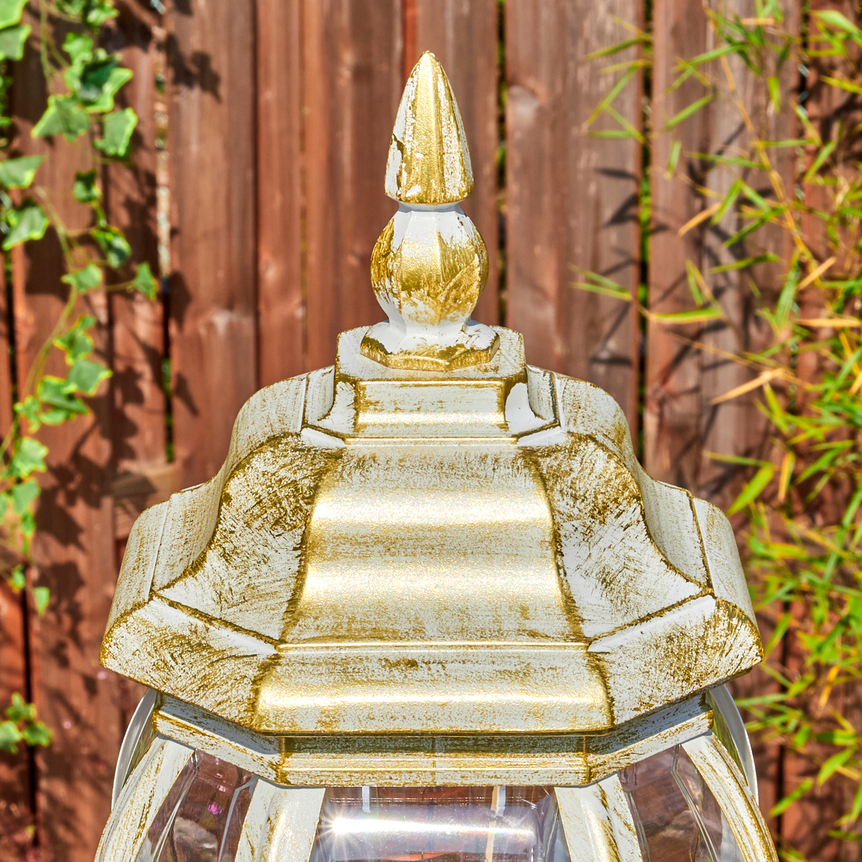 Nahaufnahme einer dekorativen Lampe mit goldenen und weißen Details. Der Hintergrund zeigt einen Holzzaun und grüne Pflanzen.