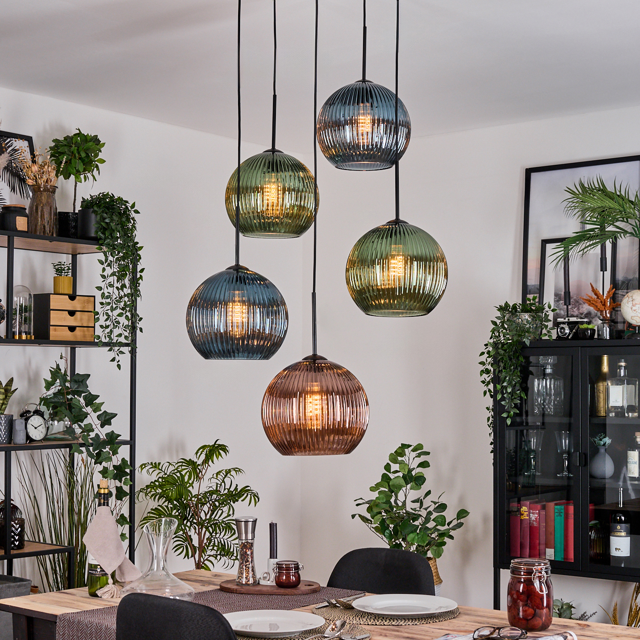 Esszimmer mit hängenden Kugelleuchten. Tisch mit Tellern und Dekoration. Regale mit Pflanzen und Dekoration. Schwarzer Schrank mit Deko.