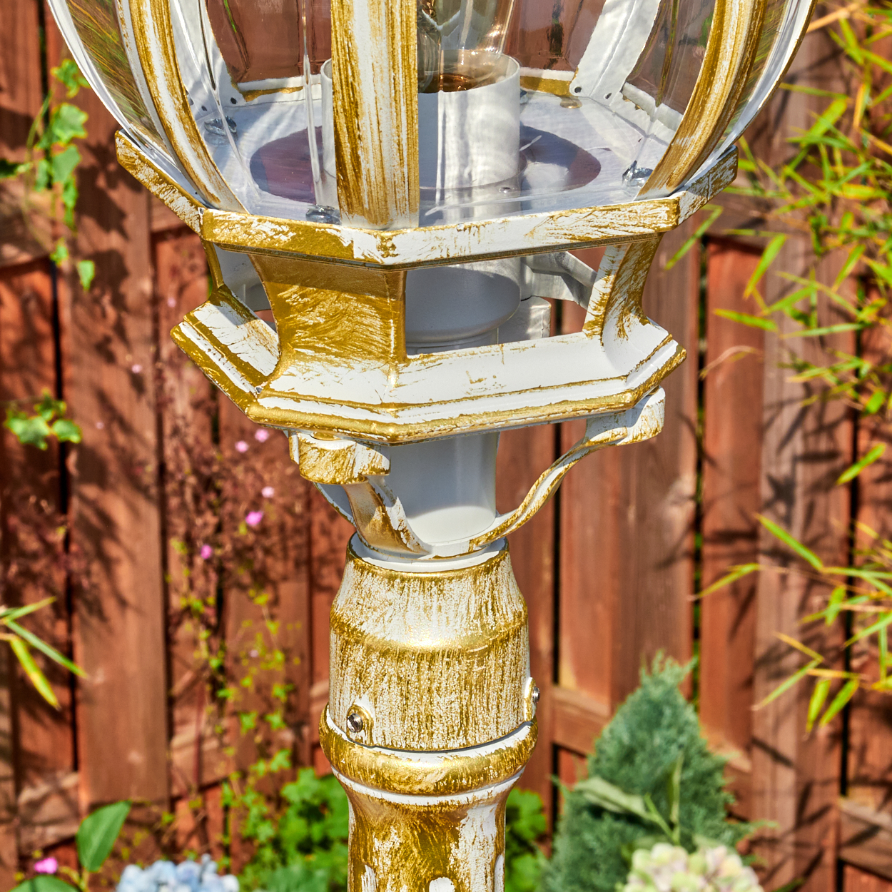 Nahaufnahme einer weißen und goldenen Außenlampe. Dahinter ein Holzzaun und Grün.