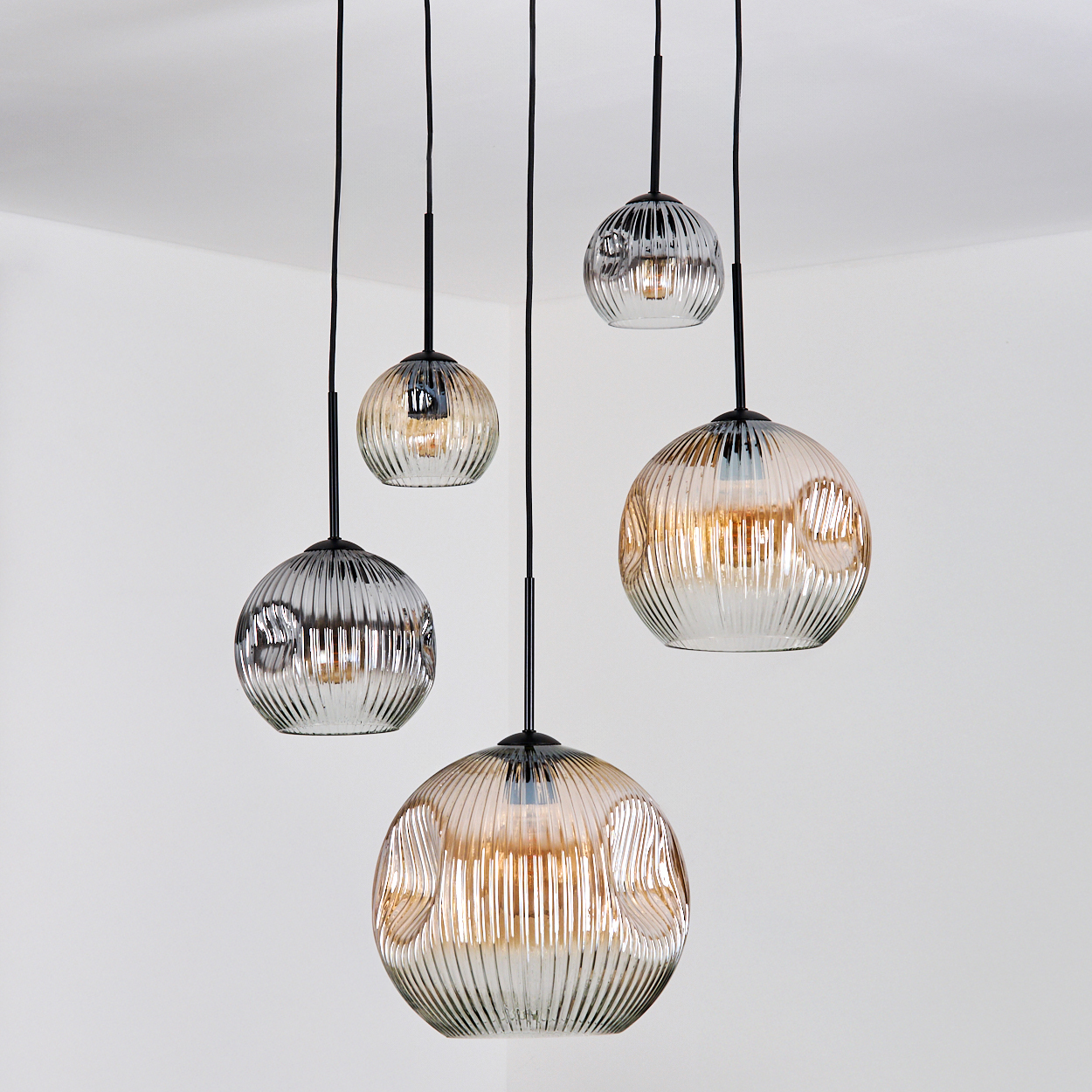 Fünf Glas-Pendelleuchten hängen von der Decke. Jede hat einen kugelförmigen, gerippten Schirm und ein schwarzes Kabel. Weiße Wände.