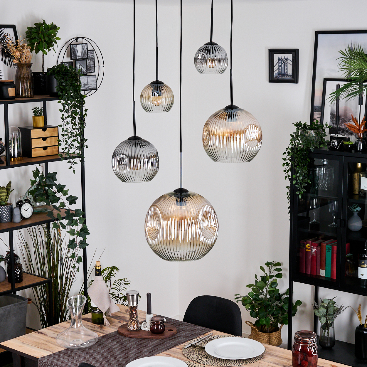 Esszimmer mit hängenden Kugelleuchten. Tisch mit Tellern, Pflanzen und schwarzen Regalen mit Dekor und Büchern. Weiße Wände.