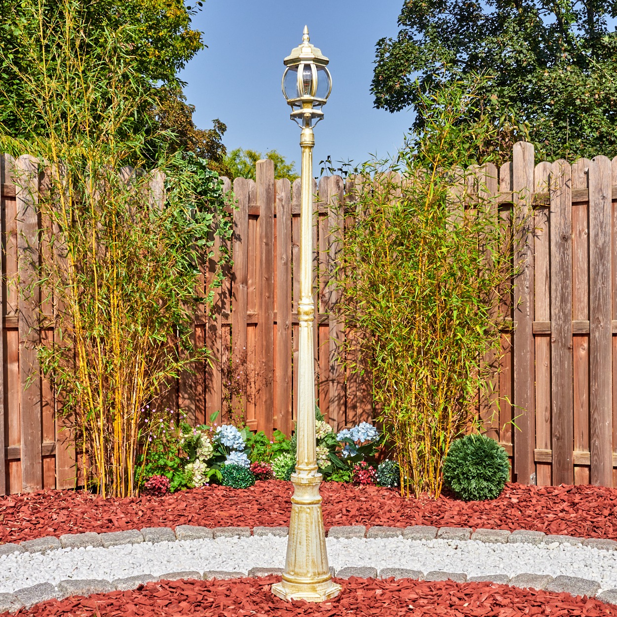 Eine goldene Laterne steht in einem Garten mit Bambus, Blumen und einem Holzzaun. Roter Mulch und ein Steinweg umgeben sie.