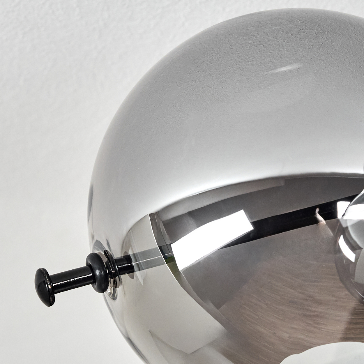 Nahaufnahme einer metallischen Kugellampe. Sie hat einen schwarzen Griff und ist teilweise reflektierend. Der Hintergrund ist eine weiße Wand.