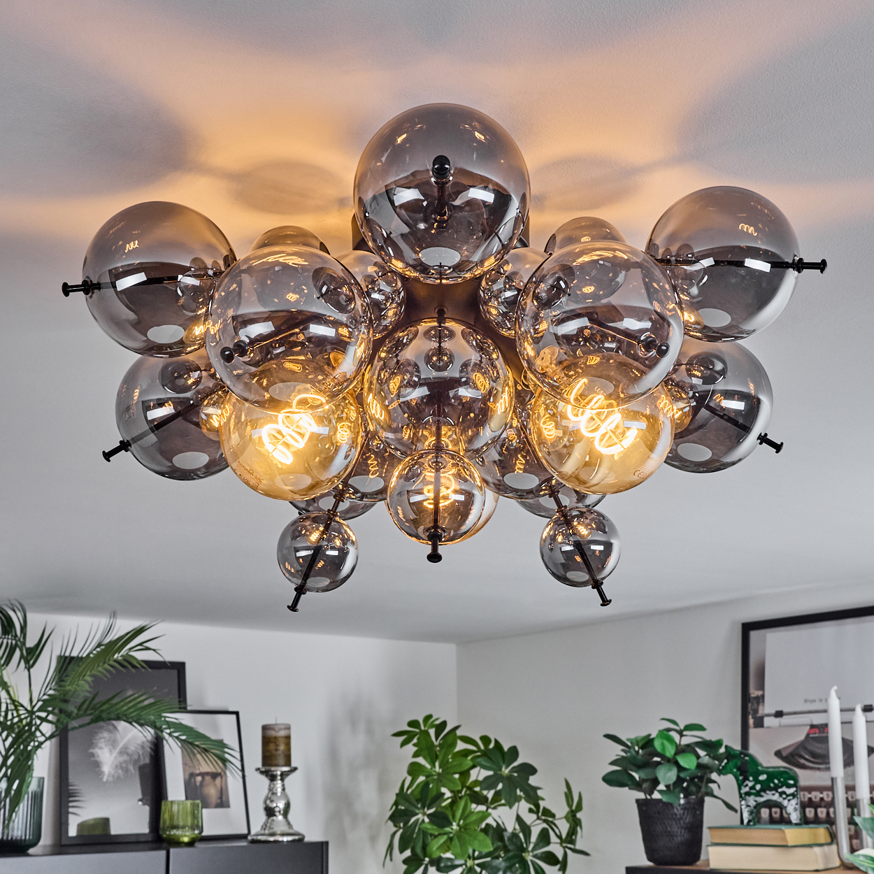 Eine moderne Deckenleuchte mit vielen Glaskugeln. Einige Kugeln sind mit warmem Licht beleuchtet. Pflanzen und gerahmte Bilder sind unten sichtbar.