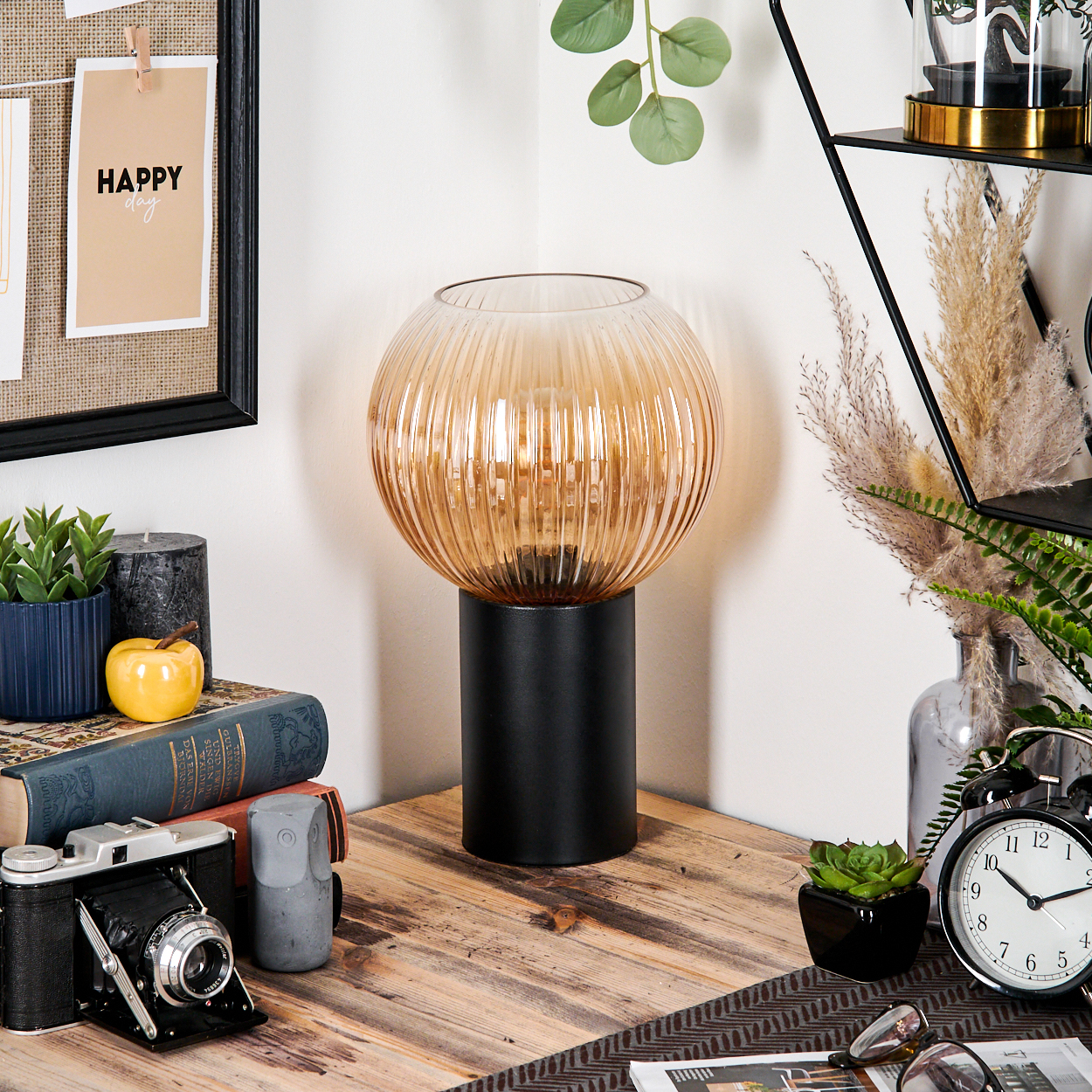 Eine dekorative Szene mit einer Lampe, Büchern und anderen Gegenständen. Eine 'HAPPY'-Notiz ist links angepinnt. Eine Kamera steht auf der Holzoberfläche.