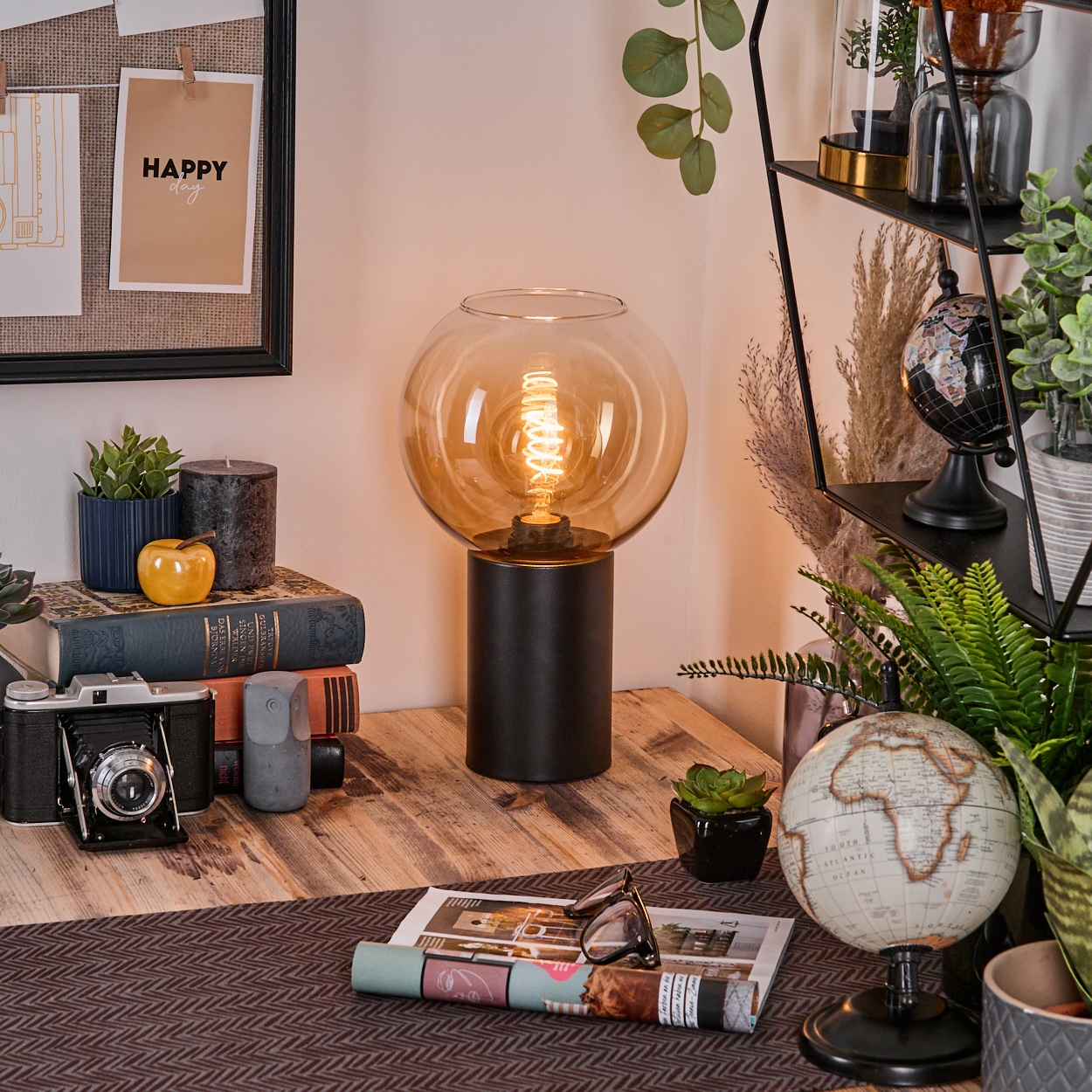 Ein Schreibtisch mit Lampe, Kamera, Büchern und Globus. Eine Pinnwand mit einer fröhlichen Notiz ist im Hintergrund.