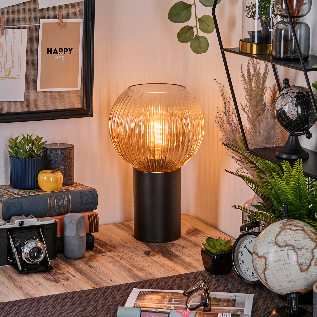 Eine leuchtende Lampe steht auf einem Holzschreibtisch. Daneben Bücher, eine Kamera und ein Globus. Links hängt eine Pinnwand.