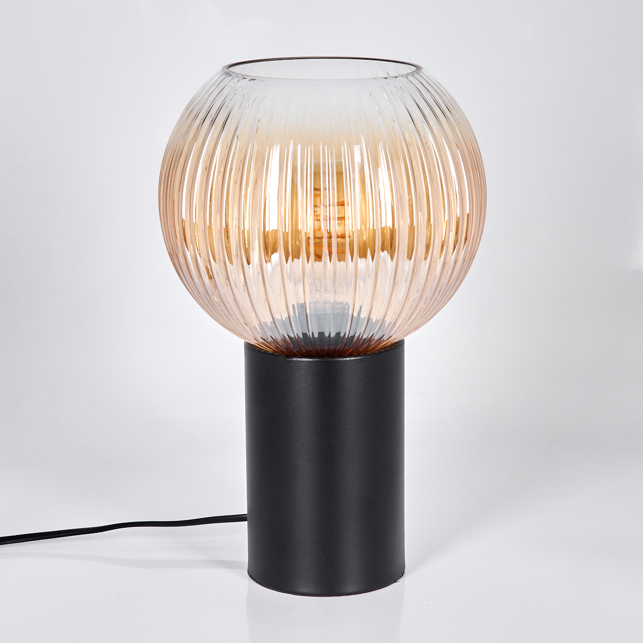 Eine moderne Lampe mit einem kugelförmigen, gerippten Glasschirm in Amber, auf einem schwarzen zylindrischen Sockel. Ein schwarzes Kabel erstreckt sich.