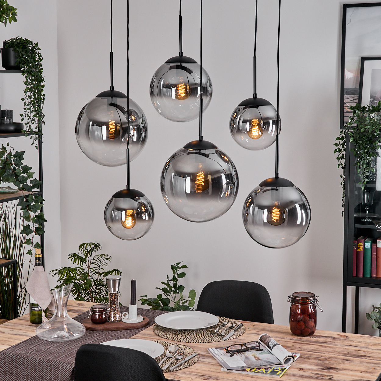 Esszimmer mit sechs hängenden Kugelleuchten, Holztisch, Pflanzen und einem schwarzen Stuhl. Verschiedenes Esszubehör liegt auf dem Tisch.