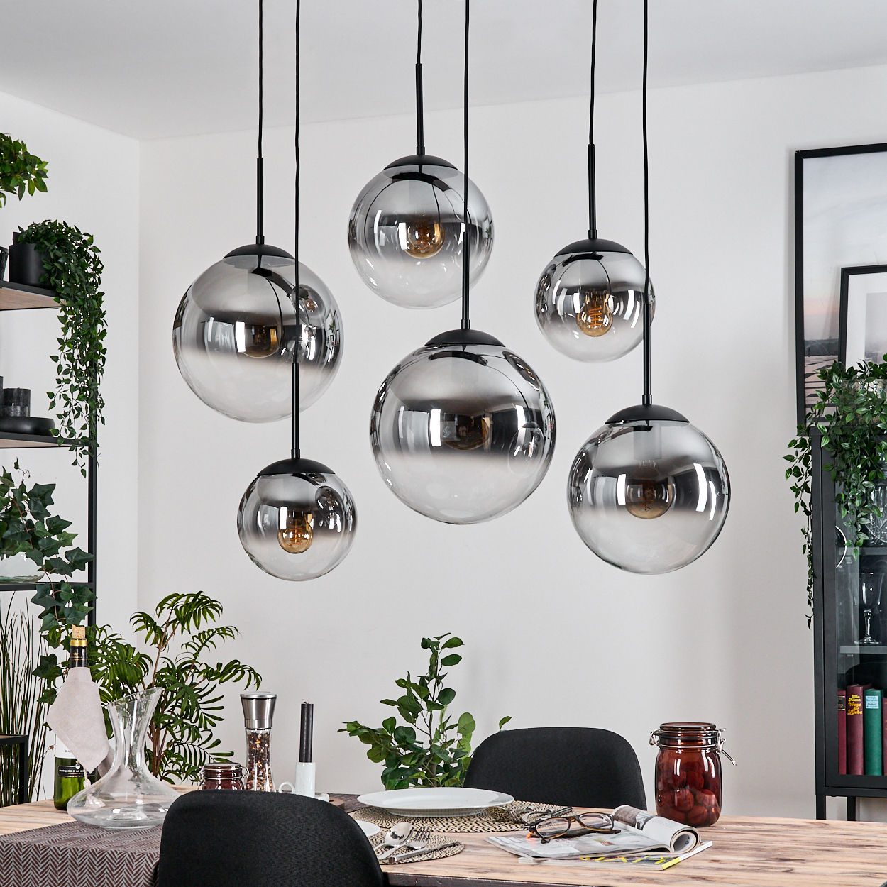 Esszimmer mit hängenden Kugelleuchten, Tisch mit Tellern, Gläsern und Essen. Pflanzen und ein Regal sind im Hintergrund.
