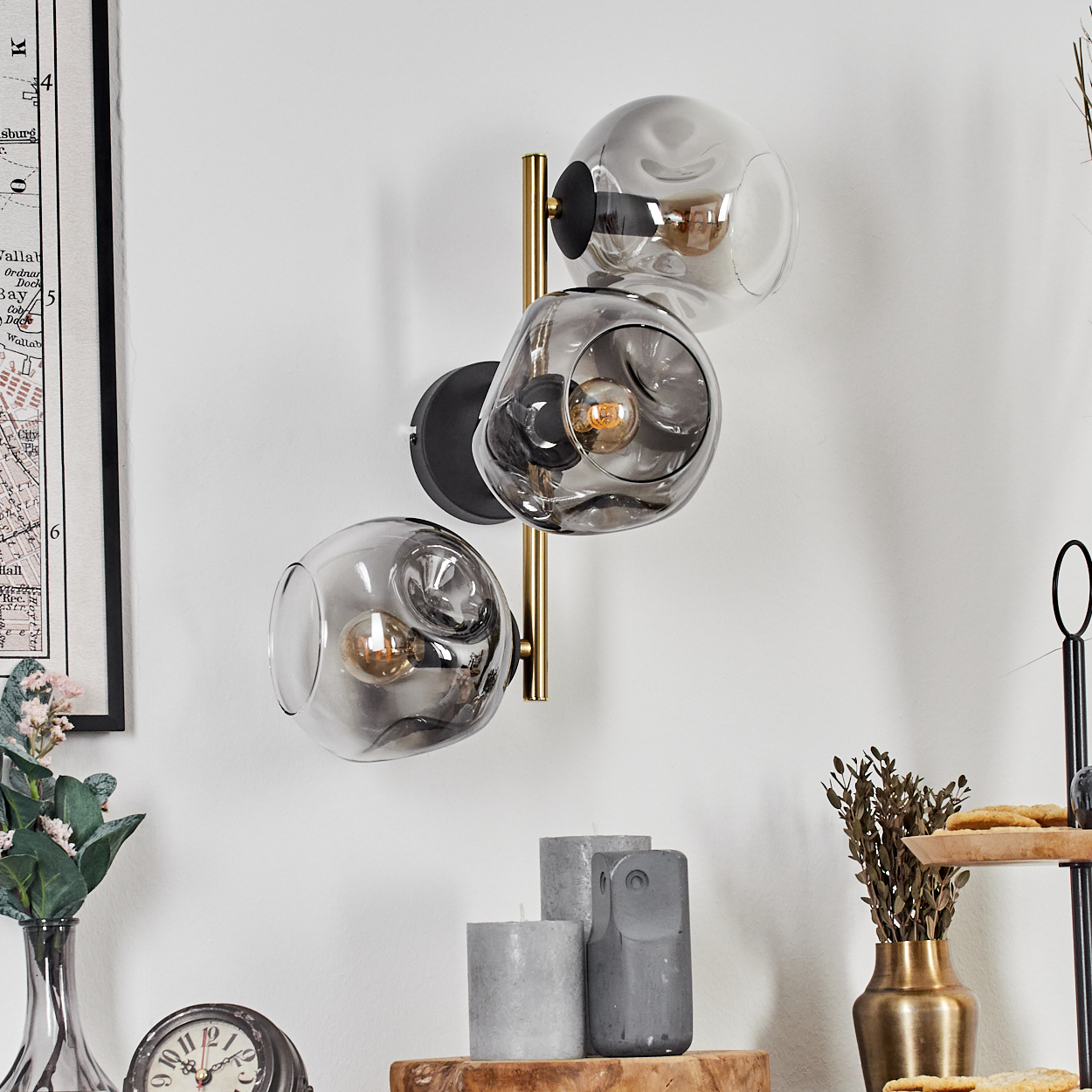 Moderne Wandlampe mit drei Glaskugeln. Kerzen, Uhr, Vase und Karte im Vordergrund.