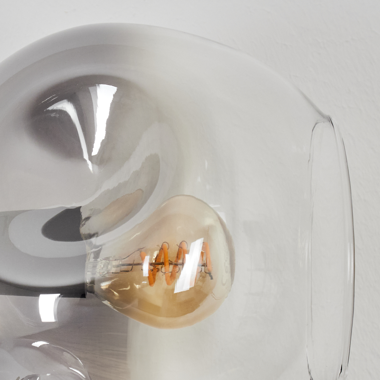 Nahaufnahme einer Leuchte mit einem transparenten Glasschirm und einer sichtbaren Glühbirne. Die Leuchte befindet sich vor einer weißen Wand.