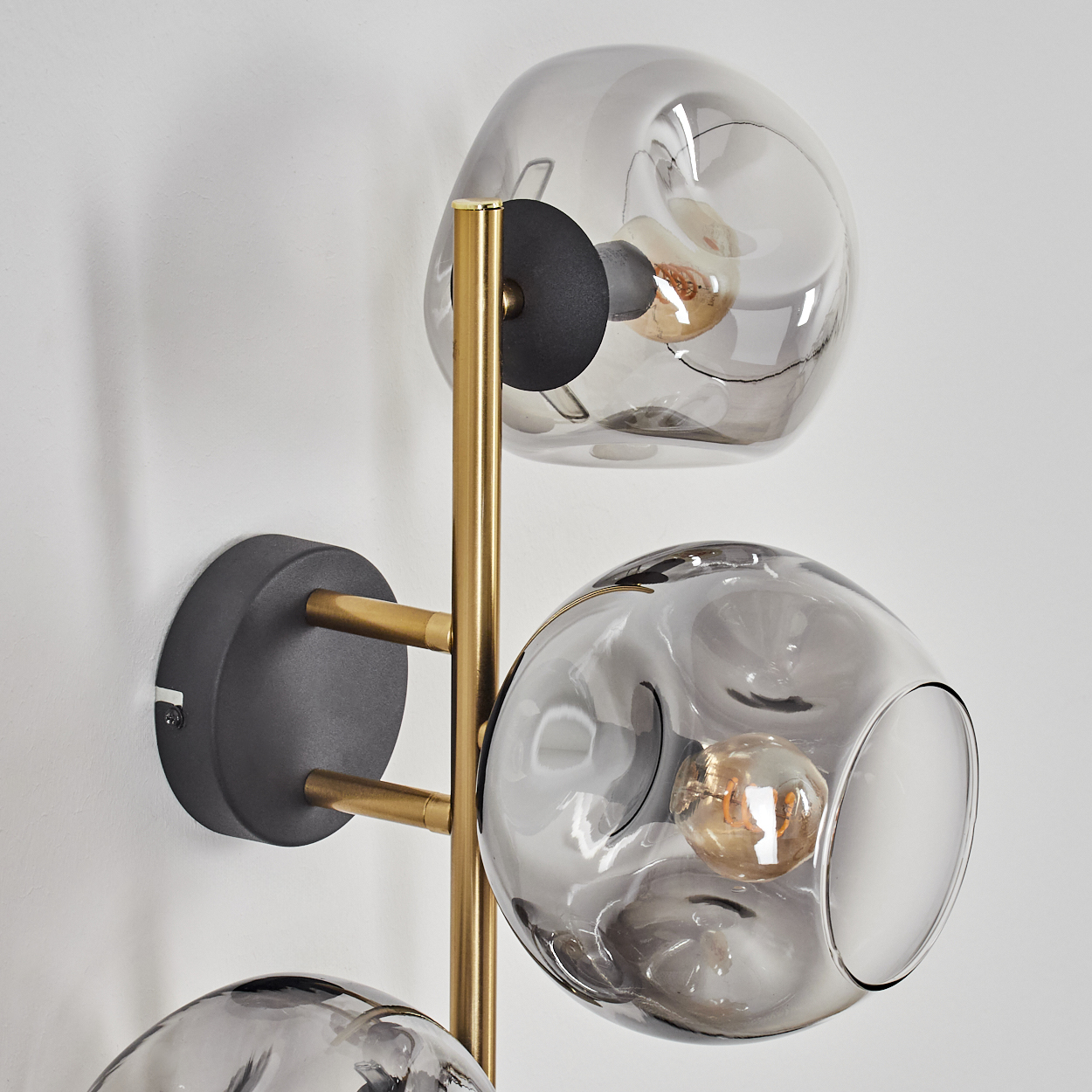Eine moderne Wandlampe mit drei Glaskugeln. Goldfarbener Metallrahmen, grauer Sockel, Glühbirnen in den Kugeln und weißer Wandhintergrund.