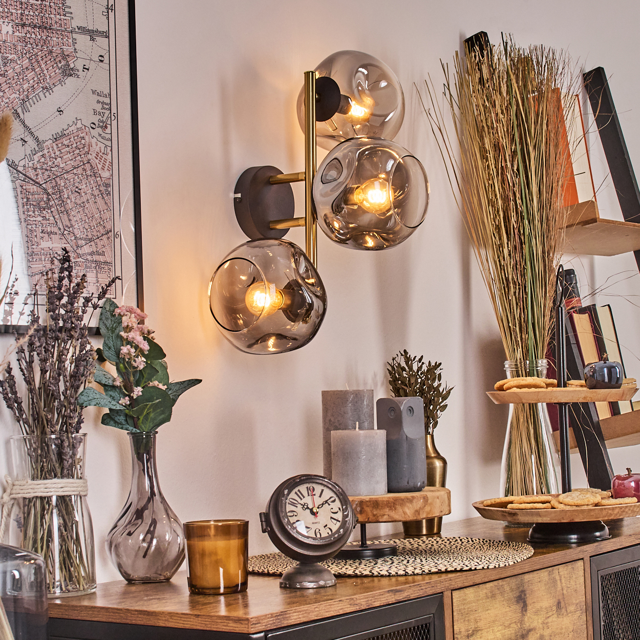 Ein Raum mit Einrichtung. Eine Wandlampe mit drei Glaskugeln, eine Vintage-Uhr, Kerzen und ein Kartenbild sind sichtbar.