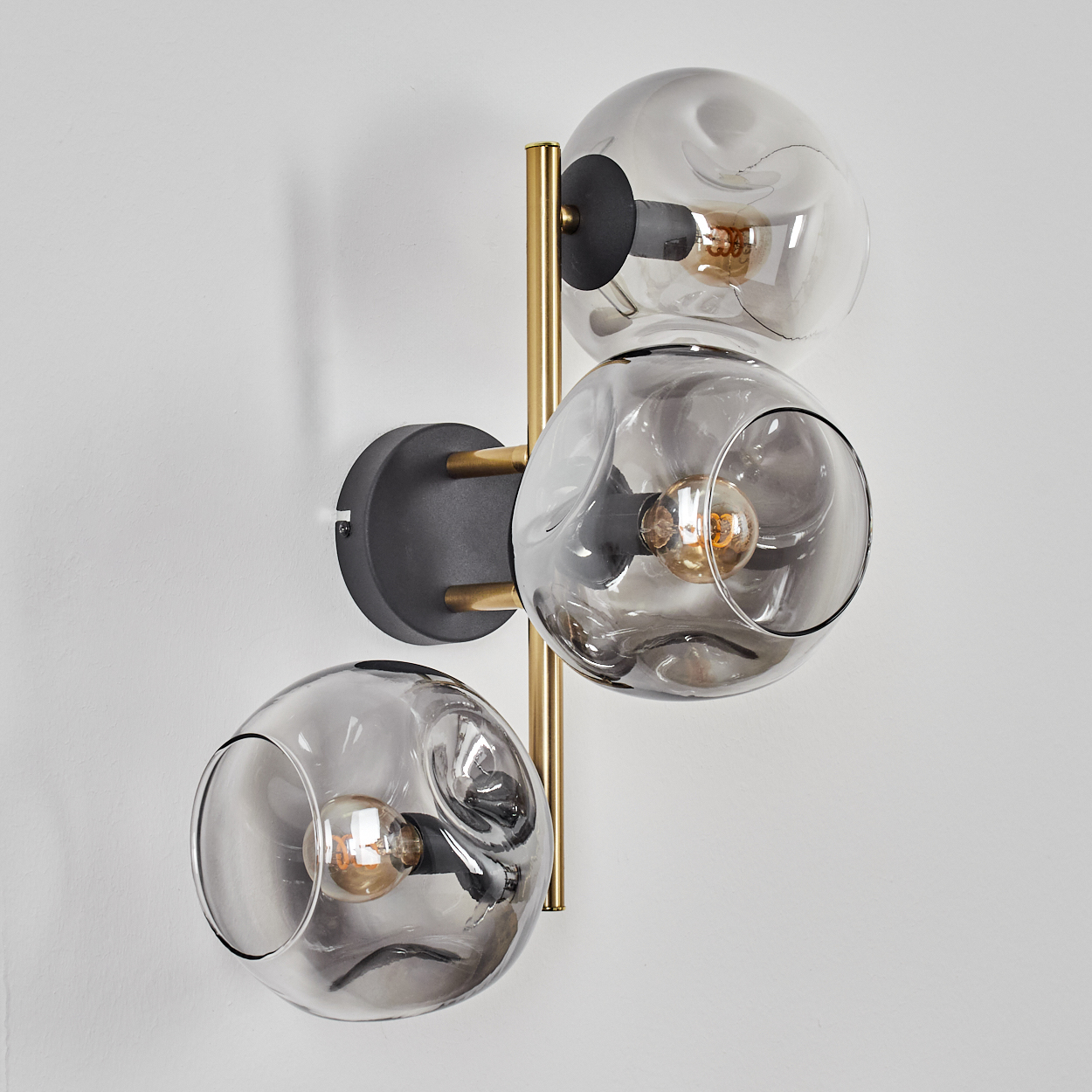Eine moderne Wandlampe mit drei Glaskugeln, die jeweils eine Glühbirne enthalten. Die Lampe hat einen gold-schwarzen Metallrahmen, montiert an einer weißen Wand.