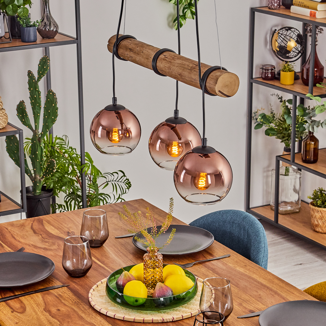 Ein Esstisch mit Obst, Gläsern und Tellern. Darüber ein Holzbalken mit drei hängenden Glaslampen. Regale mit Pflanzen.
