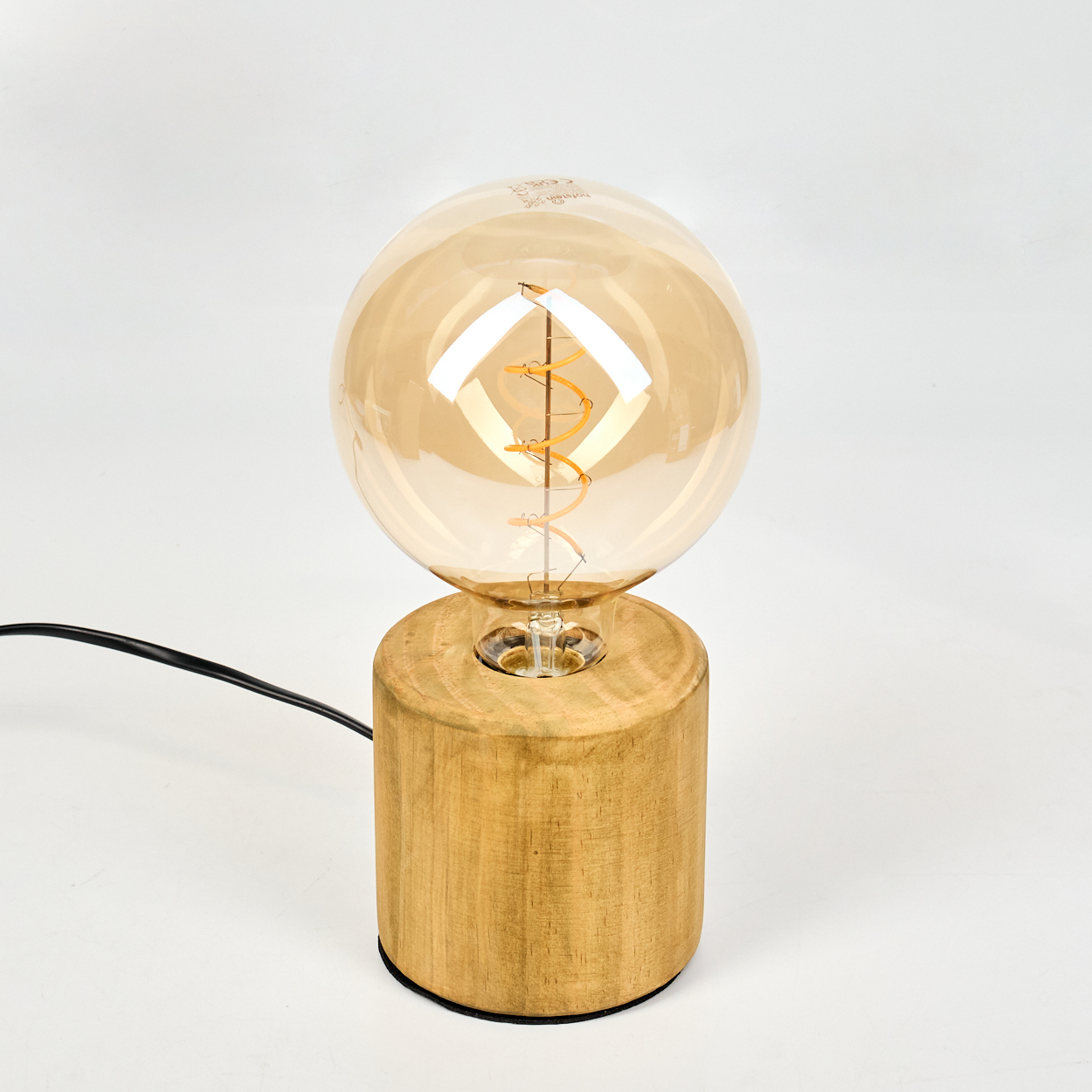Eine dekorative Lampe mit einer großen, bernsteinfarbenen Glühbirne steht auf einem Holzsockel. Ein schwarzes Kabel führt von der Basis vor weißem Hintergrund.