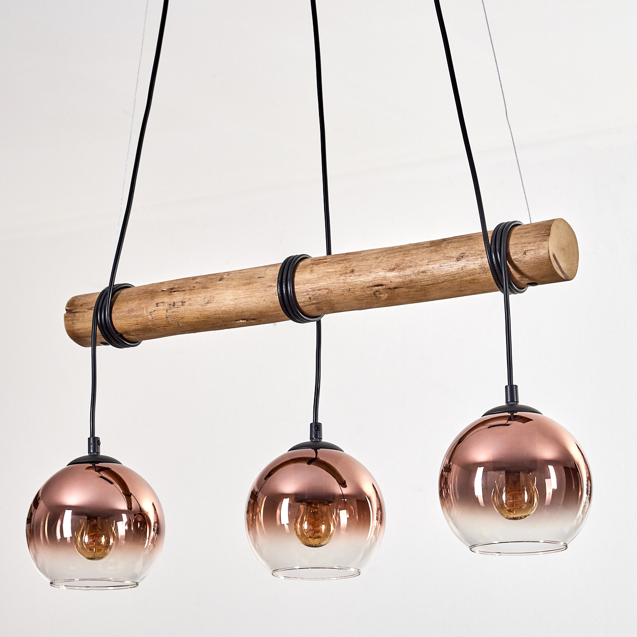 Eine moderne Hängelampe mit drei Glaskugeln. Es hat einen Holzbalken mit schwarzen Drähten und kupferfarbenen Glasschirmen.