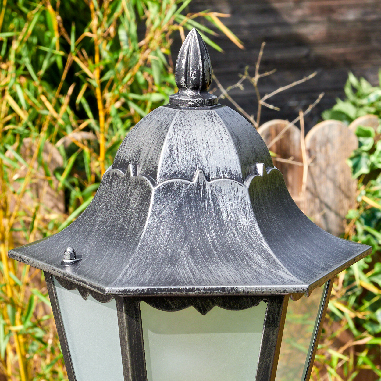 Nahaufnahme einer silbernen Außenlampe. Die Lampe hat eine dekorative Oberseite, Glasscheiben und steht vor Bambus und einem Holzzaun.