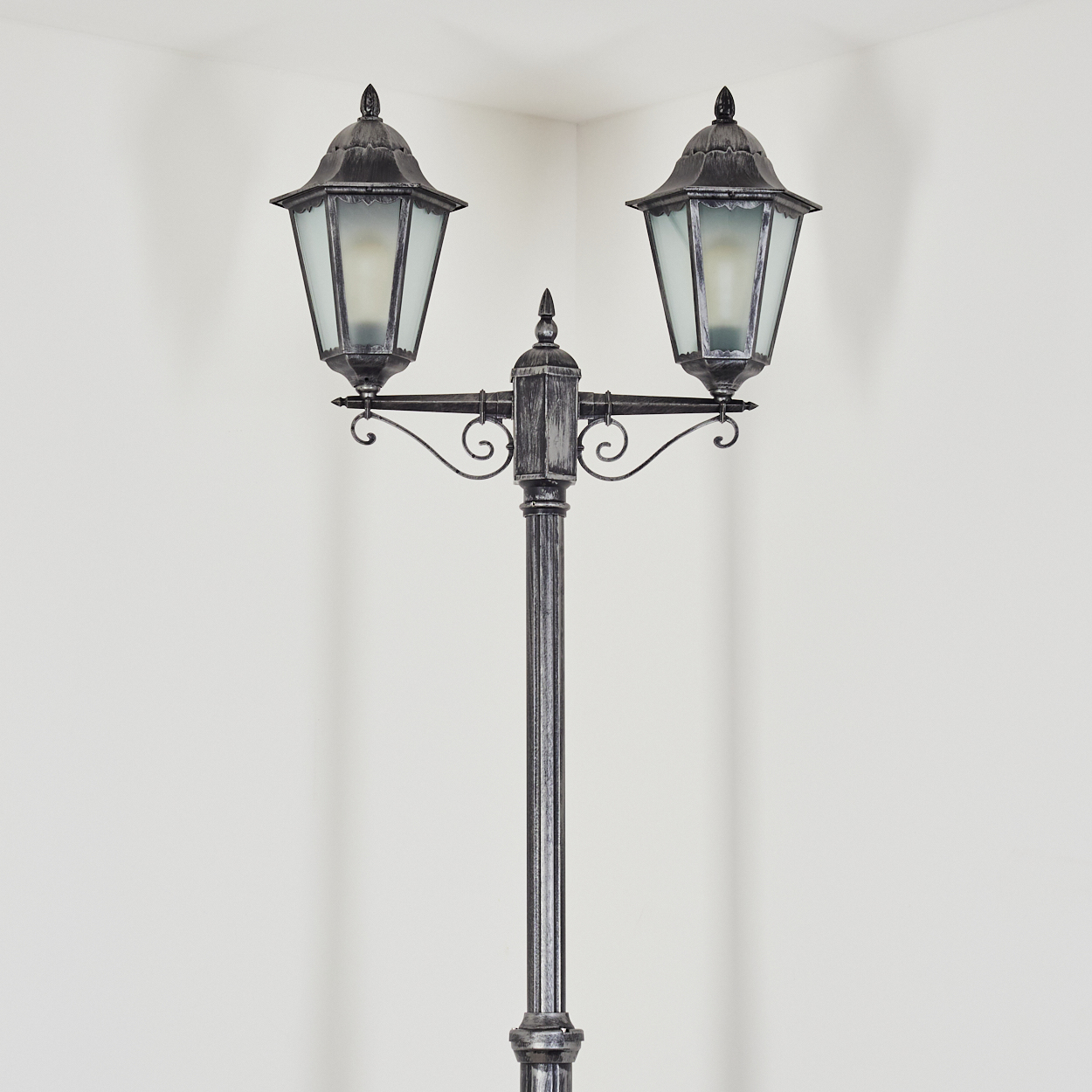 Dekorative Laterne mit zwei Laternen. Der Pfosten ist grau, die Laternen haben Glasscheiben. Der Hintergrund ist eine weiße Wand.