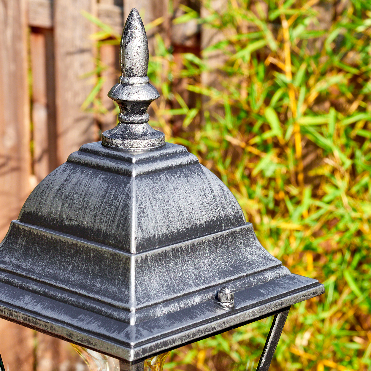 Nahaufnahme einer silbernen Außenlampe. Die Lampe hat eine spitze Oberseite. Bambus und Holzzaun sind im Hintergrund.