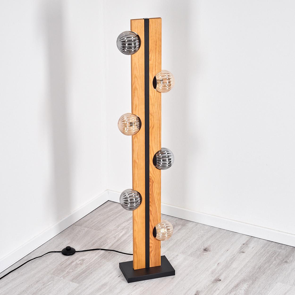 Eine moderne Stehlampe mit Holzbalken, schwarzen Akzenten und kugelförmigen Lichtern. Sie steht auf einem schwarzen Sockel vor einer weißen Wand.