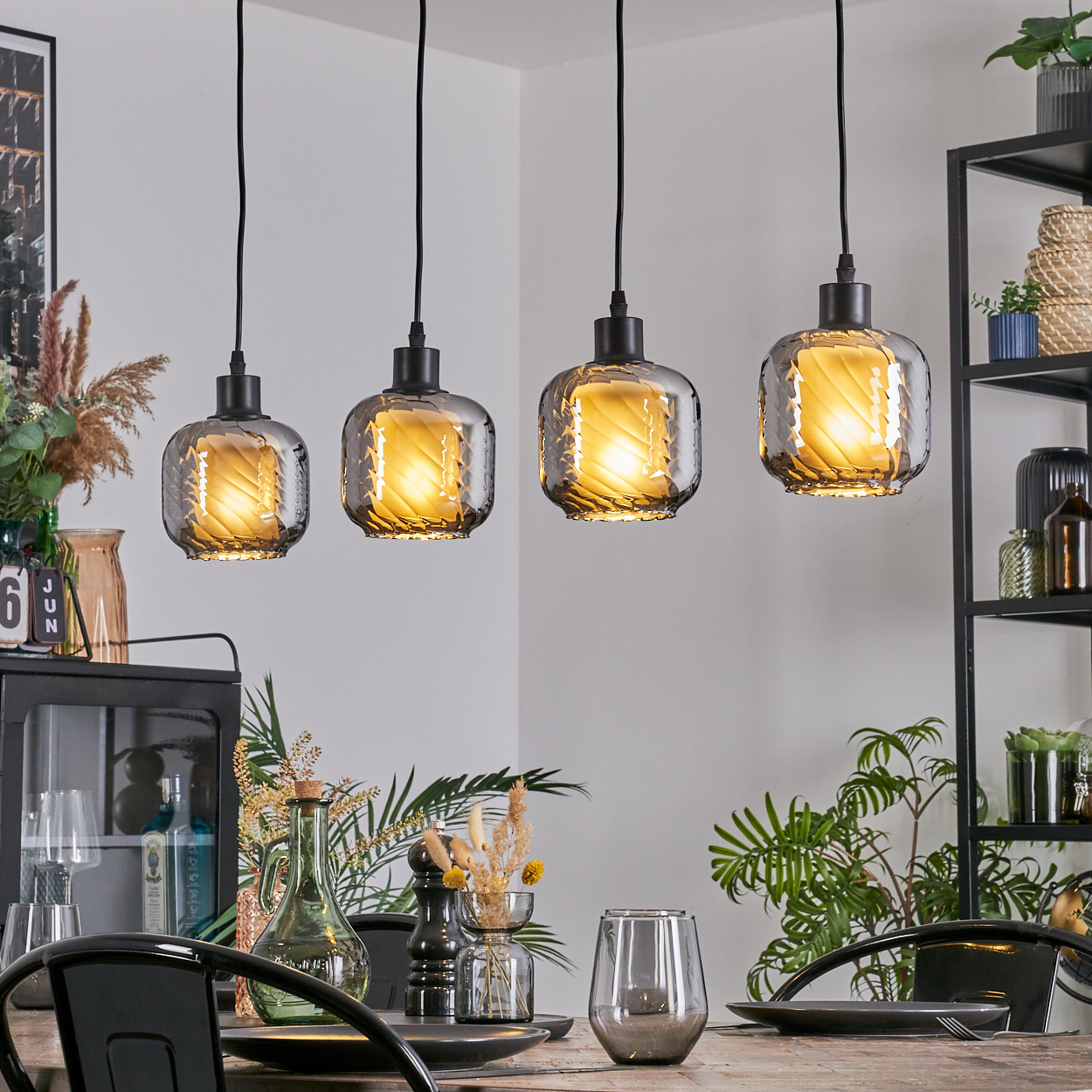 Vier Hängelampen über einem Tisch. Die Lampen sind aus Glas mit warmem Licht. Der Tisch ist mit Tellern und Gläsern gedeckt.
