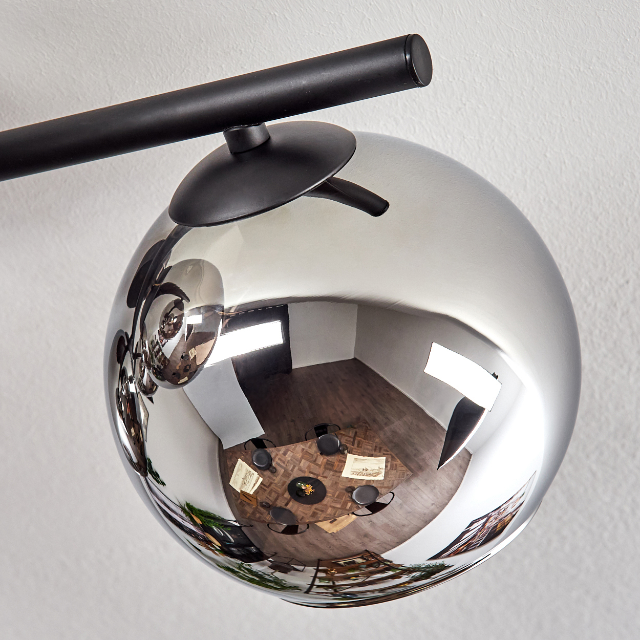 Eine reflektierende kugelförmige Leuchte. Die Kugel spiegelt einen Raum mit Tisch, Stühlen und Kunst wider. Ein schwarzer Metallarm trägt die Kugel.