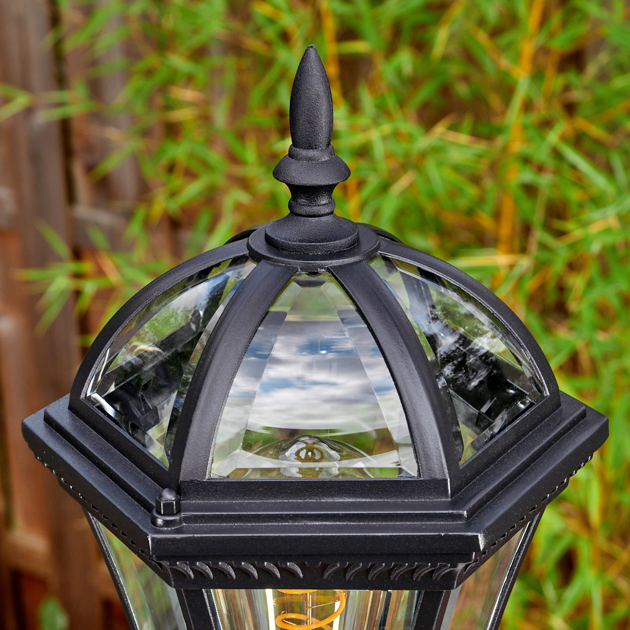 Nahaufnahme einer schwarzen Außenlampe mit Glasscheiben, vor verschwommenem grünem Bambushintergrund und Holzzaun.