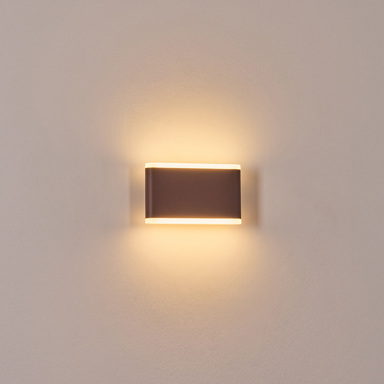 Eine rechteckige, braune Wandleuchte strahlt warmweißes Licht aus. Sie ist an einer beigefarbenen Wand angebracht.