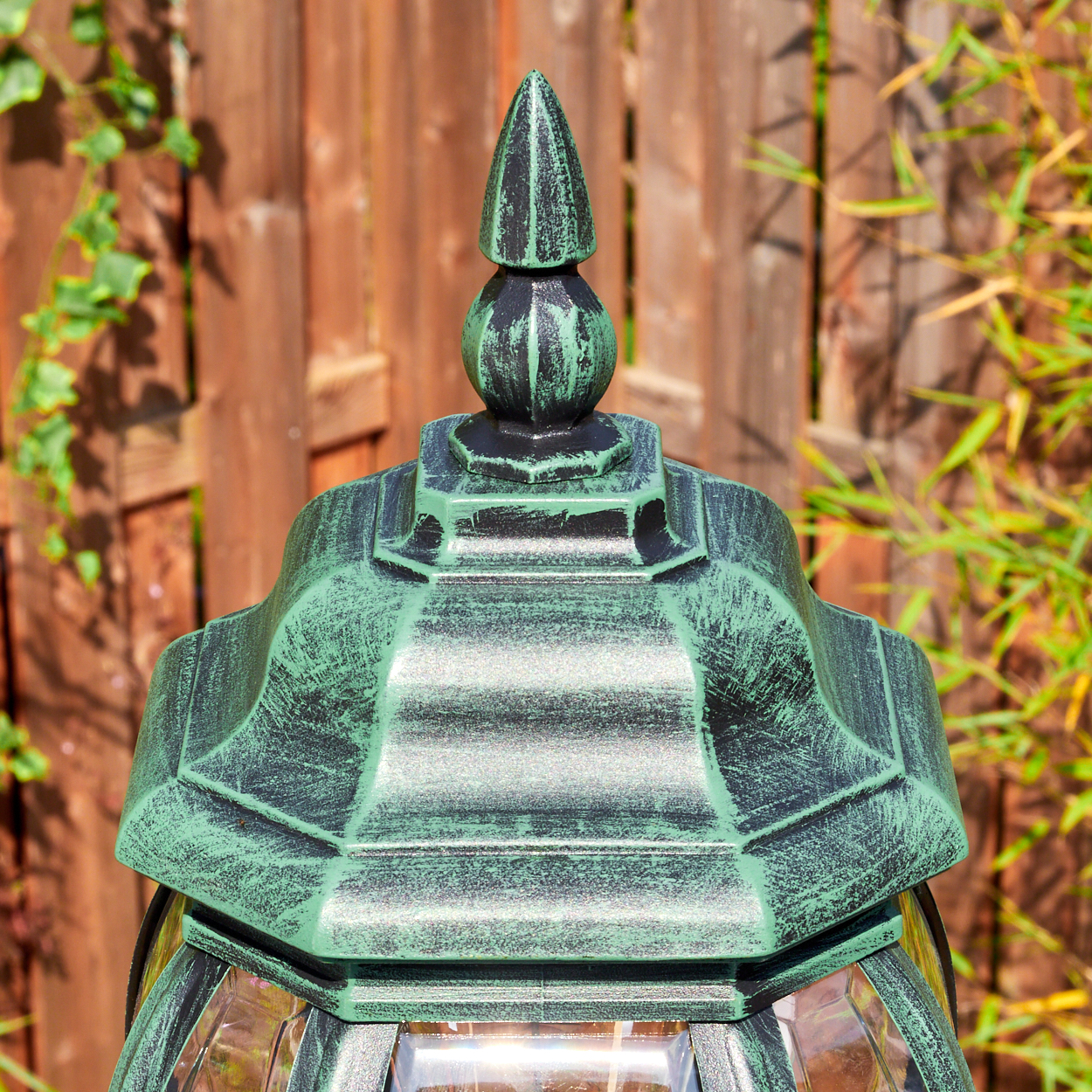 Nahaufnahme einer grün-schwarzen Außenlampe. Im Hintergrund ein Holzzaun und grüne Pflanzen.