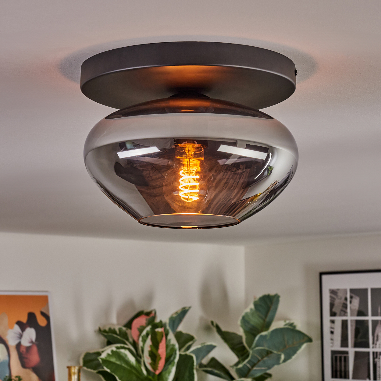 Deckenlampe mit Glasschirm und warmer Glühbirne. Pflanzen und Kunstwerke sind im unscharfen Hintergrund zu sehen.
