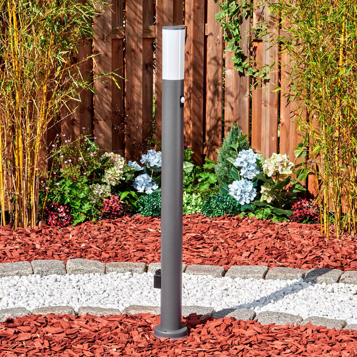 Eine graue Laterne steht in einem Garten mit Bambus, Blumen und einem Holzzaun. Roter Mulch und weiße Steine bilden den Boden.