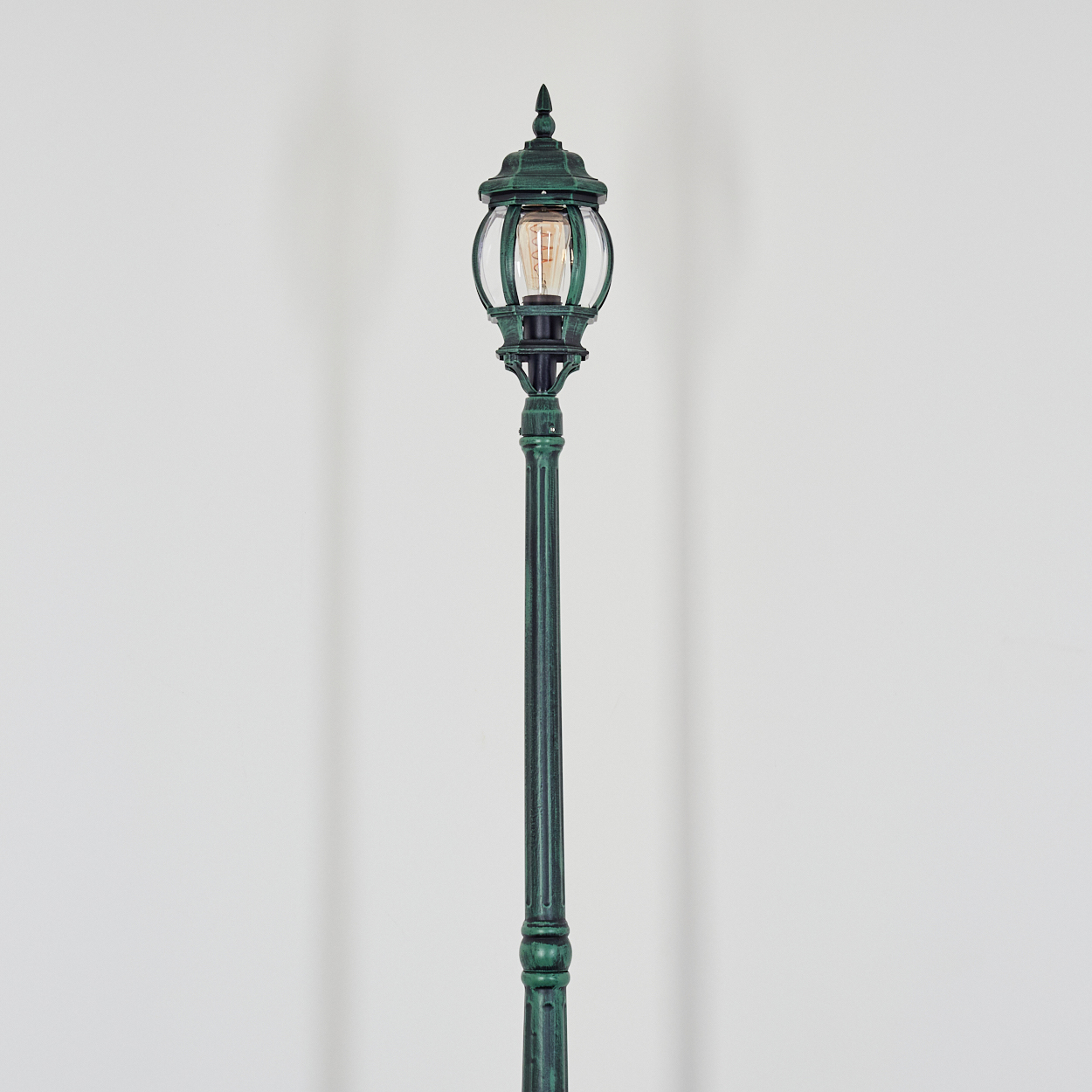 Eine grüne, verzierte Straßenlaterne steht vor einem weißen Hintergrund. Die Lampe hat eine Glaskugel, eine Glühbirne und eine spitze Spitze.