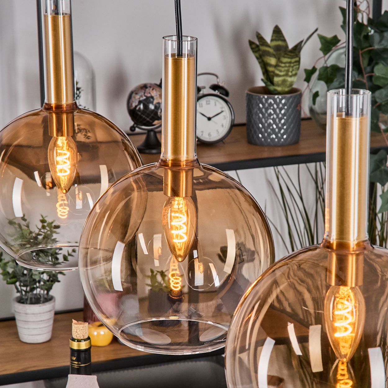 Drei Pendelleuchten mit bernsteinfarbenen Glasschirmen und goldenen Akzenten hängen. Im Hintergrund befindet sich ein Regal mit Dekoration.