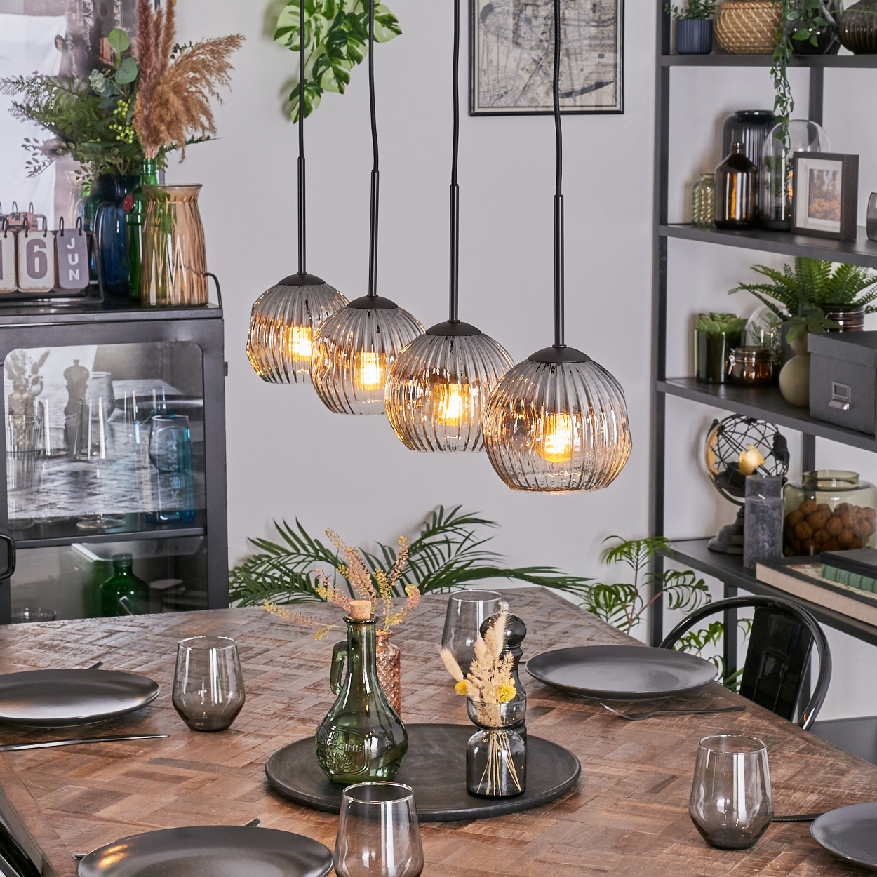 Esszimmer mit Holztisch, Glas-Pendelleuchten und Dekoration. Teller, Gläser und Blumen stehen auf dem Tisch.