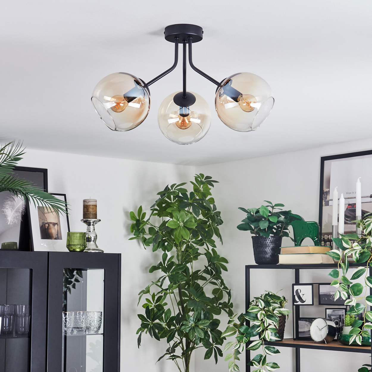 Eine moderne Deckenlampe mit drei bernsteinfarbenen Kugeln. Der Raum hat Pflanzen, einen schwarzen Schrank, Regale mit Dekor und eine weiße Decke.